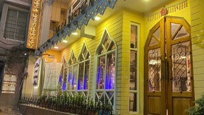 Um edifício com uma placa indicando 'Omega Residency', apresentando uma varanda com guarda-corpo, uma série de janelas com vidro decorativo e uma porta com portão de metal.