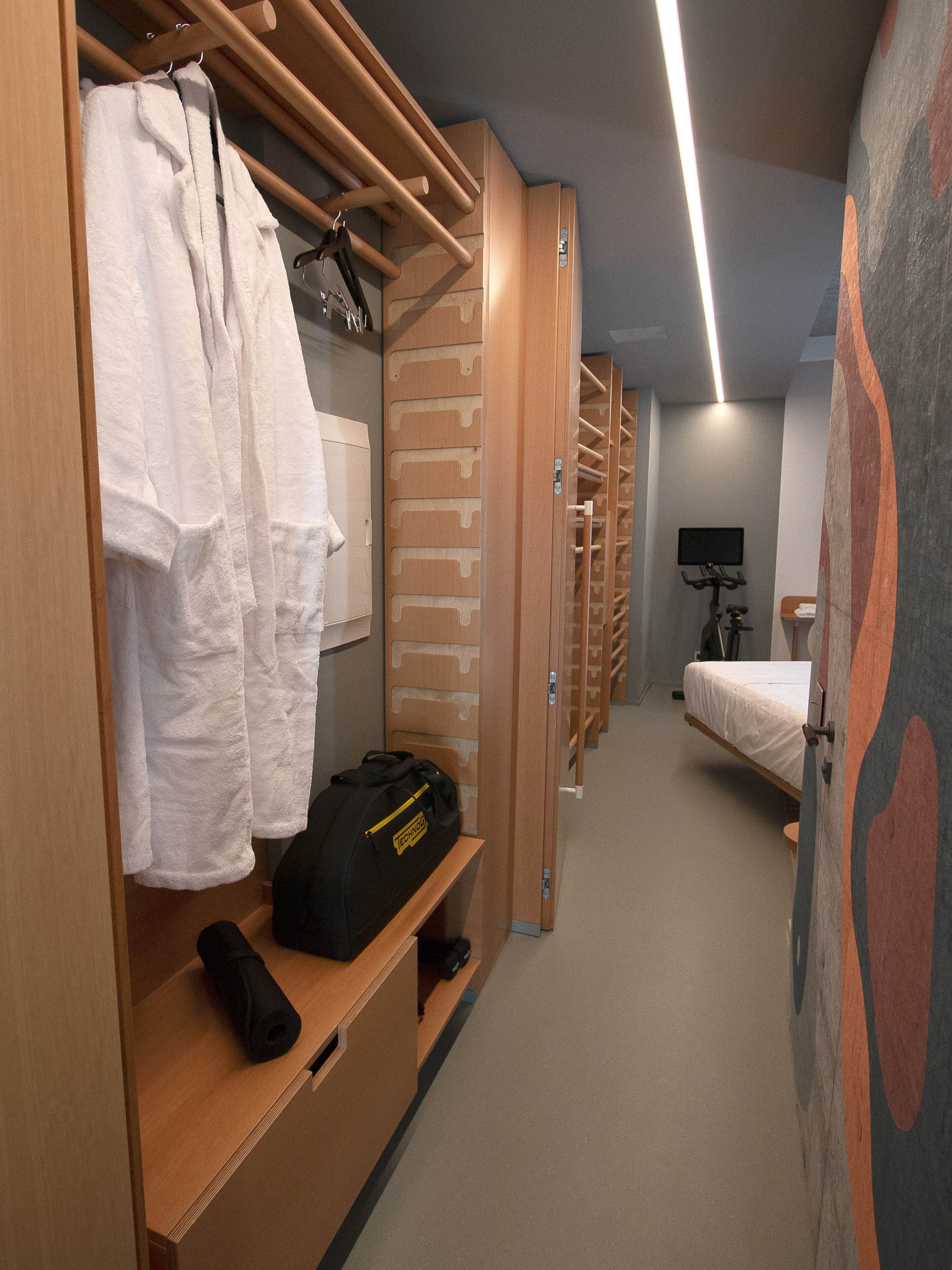 A hotel room with a wooden wardrobe, a bed, a desk with a bag, and a TV.
