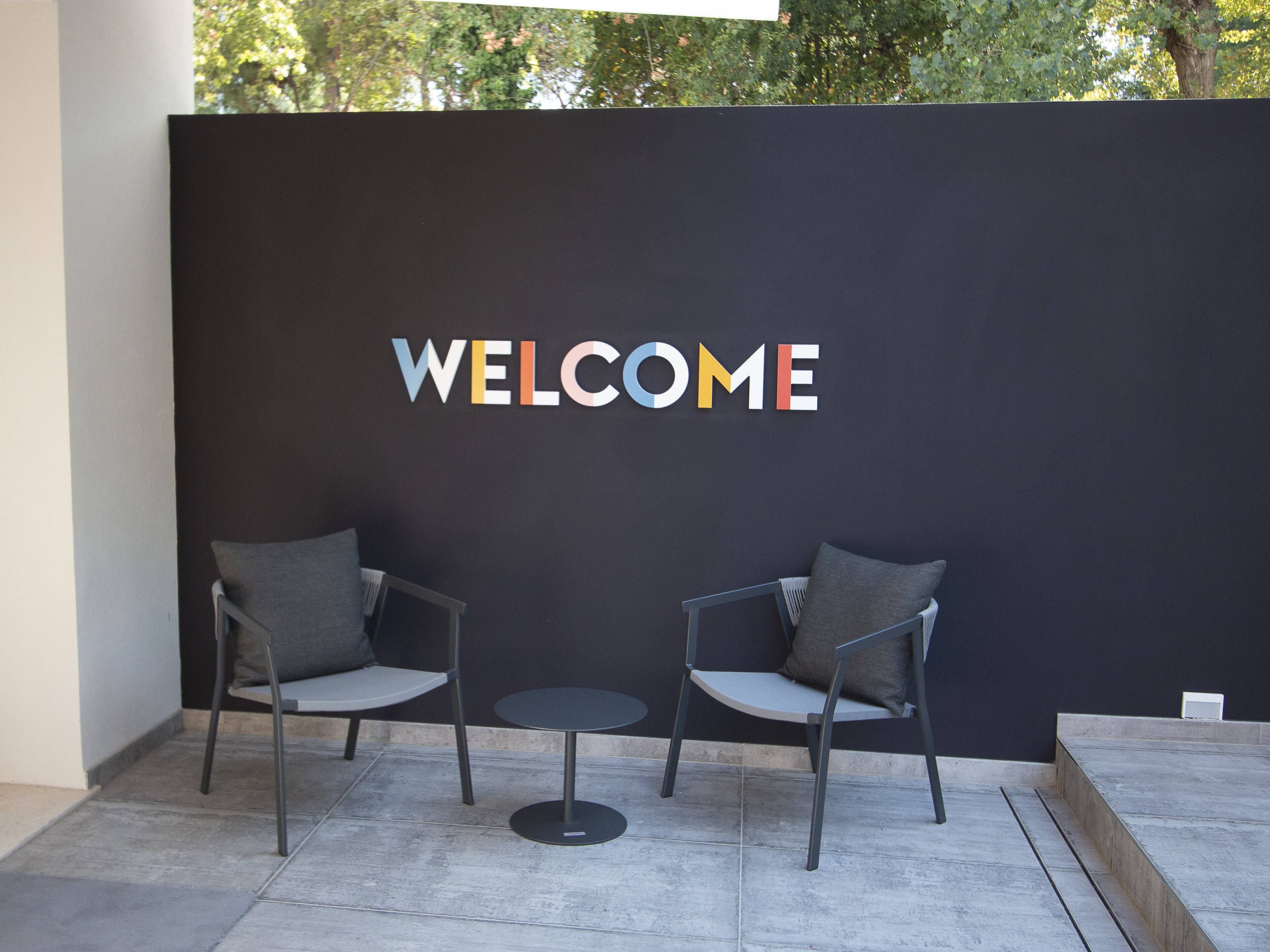 Un espace d’accueil avec une pancarte indiquant « BIENVENUE », deux fauteuils gris et une petite table ronde.