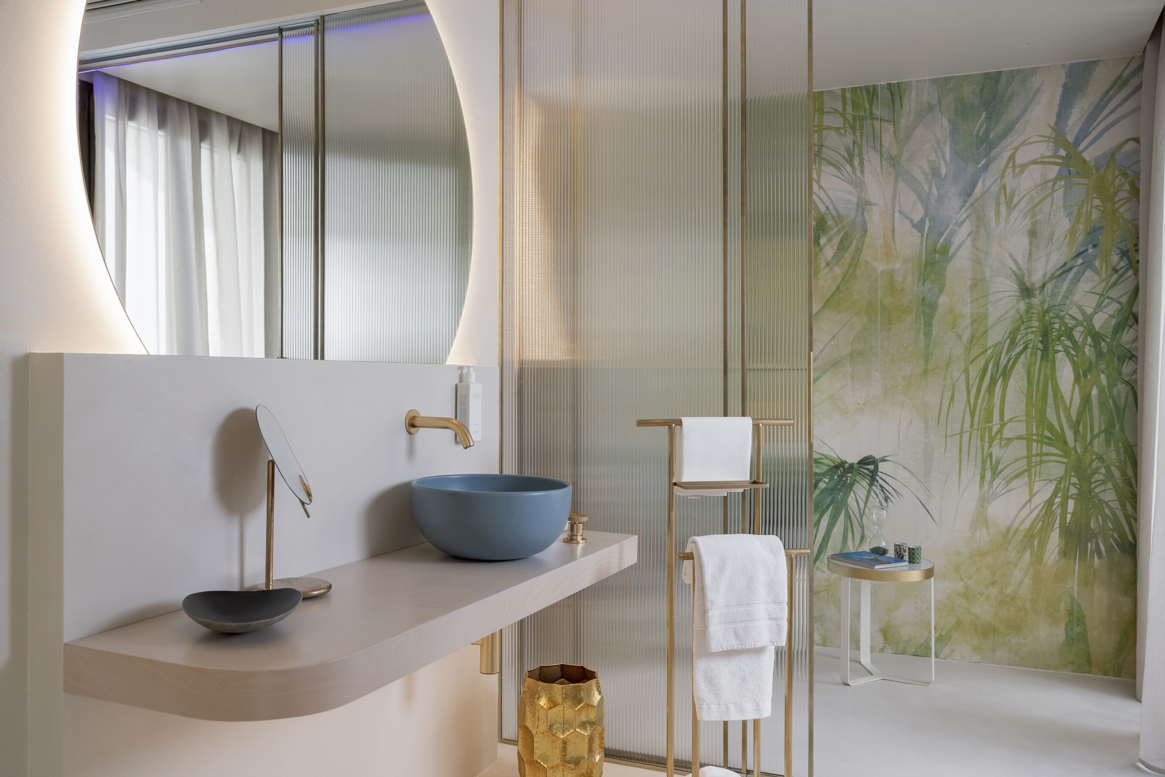 A modern bathroom with a large mirror, a blue vessel sink, a gold vase, and a tropical mural.