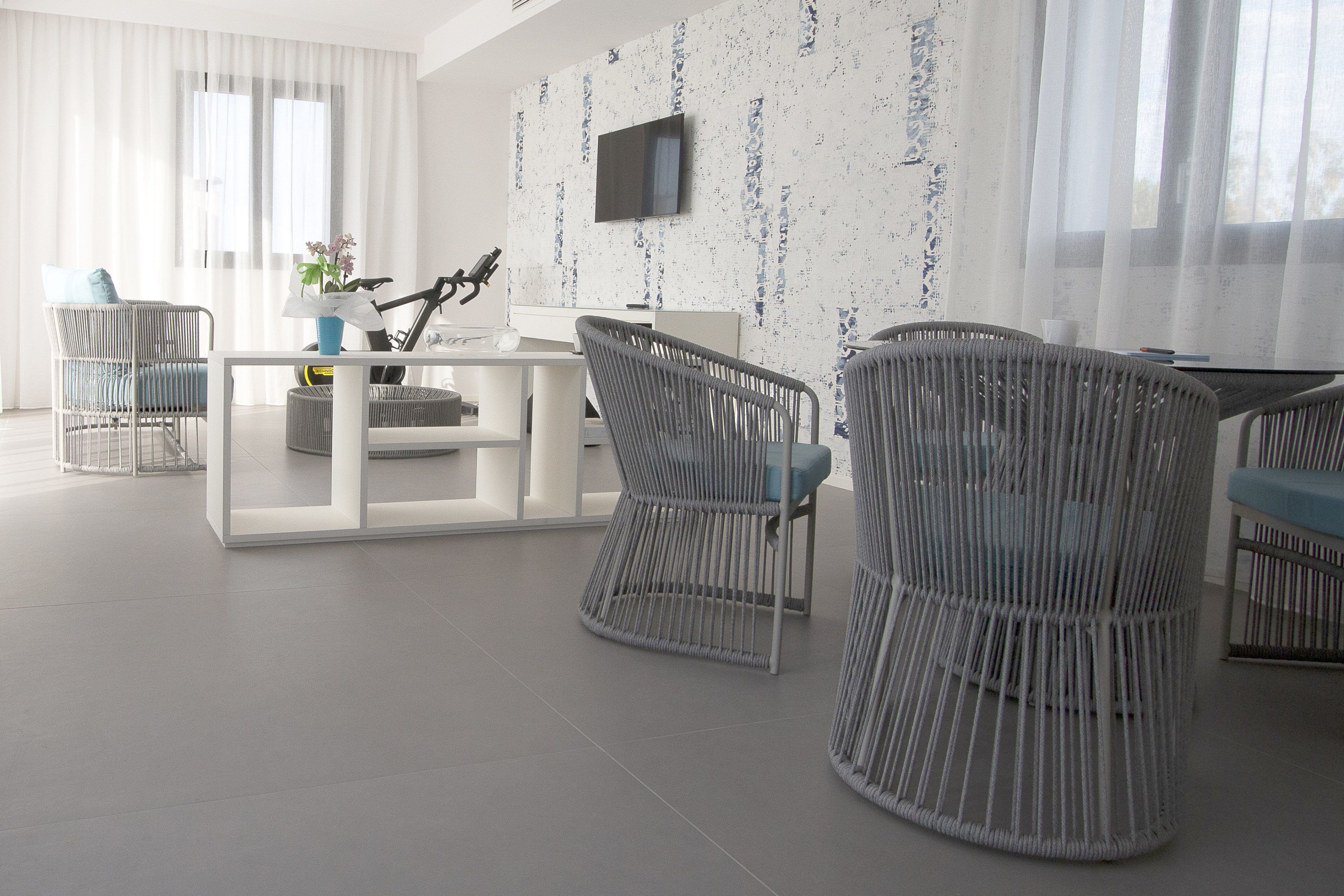 A modern, well-lit room with a white sofa, grey wicker chairs, a white coffee table, and a flat-screen TV mounted on the wall.