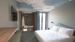 A hotel room with a bed, a wooden desk, a mirror, a bathroom with a towel rack, and a ceiling with a floral design. - In A Light Wave: Il mare, l’acqua. Flexibility and inclusiveness (Rimini)