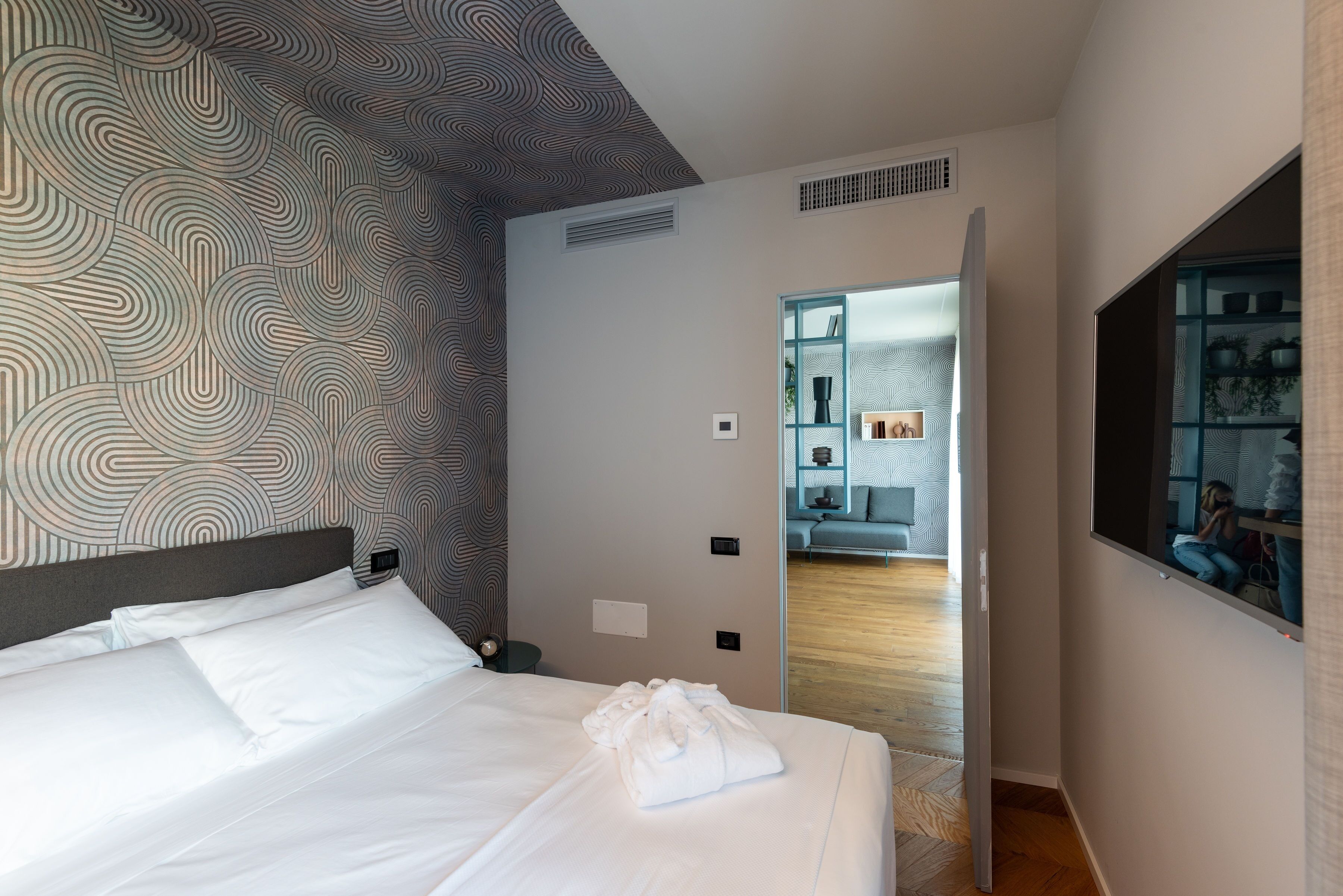 A modern hotel room with a large bed, a patterned wall, and a view into the living area.