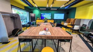 Un salón de conferencias con una mesa de madera, sillas amarillas y verdes, una pantalla de proyector y un ventanal grande.