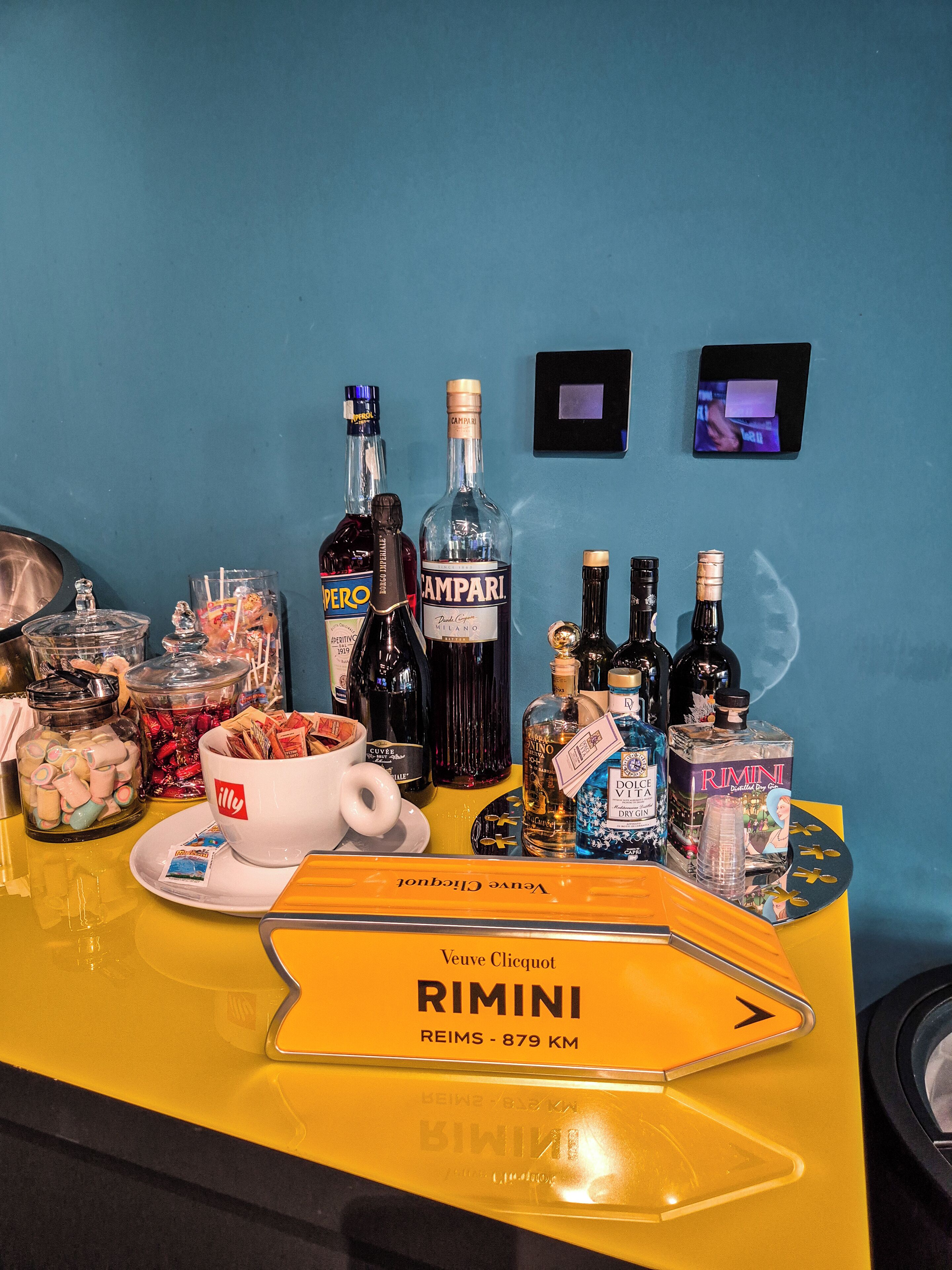 Eine Bar mit verschiedenen alkoholischen Getränken, einer Kaffeetasse und einem Tablett mit der Aufschrift "Veuve Clicquot RIMINI REIMS - 879 KM".