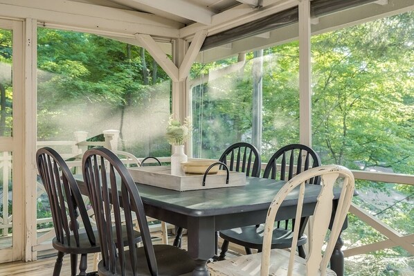 Une terrasse couverte, dotée d’une table et de chaises, est entourée de baies vitrées qui offrent une vue sur une zone boisée.