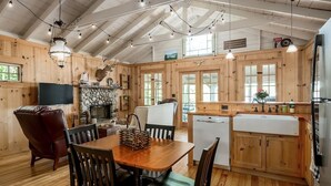 A rustic cabin interior with a wooden dining table, chairs, a fireplace, a kitchen area with a sink and cabinets, and a television. - Dune-Top Harbor Views / Big Red Cottage + Hot Tub / Treetops (Saugatuck)
