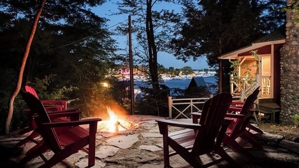 A stone deck with Adirondack chairs and a fire pit, overlooking a coastal view. - Dune-Top Harbor Views / Big Red Cottage + Hot Tub / Treetops (Saugatuck)