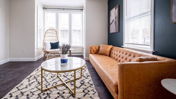 A living room with a round coffee table, a sofa, a wicker chair, and a large window with blinds. - ★ Modern Renovation ★ 10-Bedrooms! Perfect For Large Groups! (Providence)