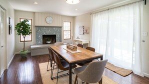 Un comedor con mesa de madera, sillas, chimenea y un amplio ventanal con cortinas translúcidas.