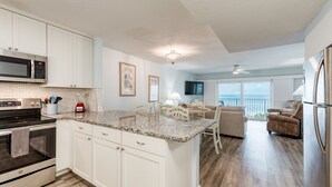 A modern kitchen with white cabinets, a granite countertop, stainless steel appliances, and a dining area with a view of the ocean. - Direct Ocean- Wrap Around Balcony (Indialantic)
