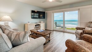 A living room with a television, a sofa, a coffee table, and a view of the ocean through large sliding glass doors. - Direct Ocean- Wrap Around Balcony (Indialantic)