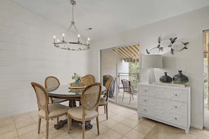 Un coin repas avec une table ronde, quatre chaises, un lustre, un buffet blanc agrémenté d’objets décoratifs et une vue sur l’extérieur, accessible par une porte vitrée.