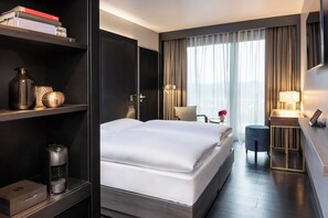 A hotel room with a large bed, a desk with a lamp, a bookshelf, and a coffee maker. - Hotel AMANO East Side (Berlin)
