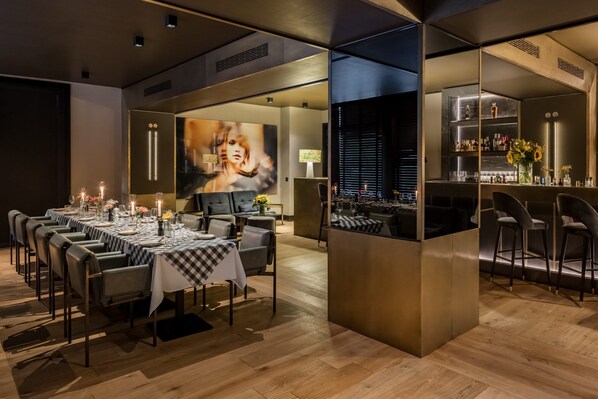 A modern dining area with tables set for a meal, a bar area with bottles and glasses, and a large screen displaying an image.