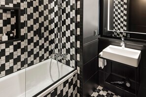 A modern bathroom with a checkered black and white tile backsplash, a white bathtub, a sink with a mirror above it, and a shower with chrome fixtures. - Hotel AMANO East Side (Berlin)