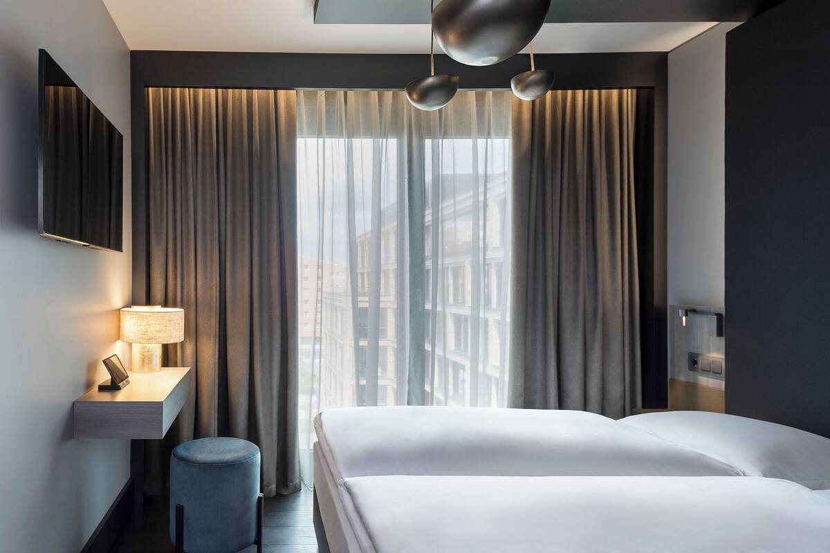 Ein modernes Hotelzimmer mit einem großen Bett, einem Nachttisch mit Lampe, einem kleinen Hocker und einem großen Fenster mit Vorhängen.