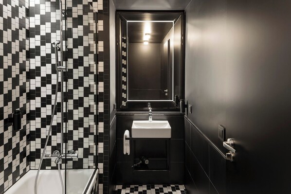 A narrow bathroom with black and white checkered tiles, a white sink, a glass shower enclosure, and a mirror above the sink. - Hotel AMANO East Side (Berlin)