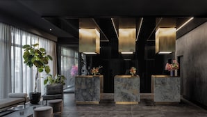 A modern reception area with marble reception desks, a seating area with a bench and ottoman, and a large potted plant. - Hotel AMANO East Side (Berlin)