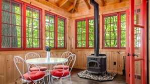Una habitación acogedora con detalles de madera, una mesa redonda con sillas, una chimenea y ventanas grandes con marcos rojos.