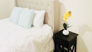 Camera da letto con testiera trapuntata, biancheria bianca, un piccolo comodino nero con un vaso di fiori gialli e un elemento decorativo a parete.
