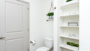 Un bagno con il water bianco, una porta bianca ed scaffali con piante e un cartello con la scritta “BELIEVE”.