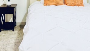 Una camera da letto con un letto, biancheria bianca, due cuscini arancioni, una testiera bianca, un elemento decorativo a parete, un piccolo tavolo con un vaso di fiori e un comodino scuro.