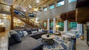 A spacious living area with a large sectional sofa, a round coffee table, and a fireplace. A staircase with wooden railings leads to an upper level. - Blue Pine Divine - 1+ Bedrooms, 2 Baths, Sleeps 6, pet friendly, Hot Tub (Broken Bow)