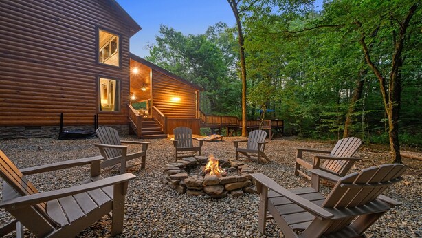 A log cabin with a fire pit surrounded by Adirondack chairs, set in a forested area. - Blue Pine Divine - 1+ Bedrooms, 2 Baths, Sleeps 6, pet friendly, Hot Tub (Broken Bow)
