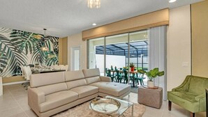 A modern living room with a beige sectional sofa, a glass coffee table, and a green armchair. There is a dining area with a table and chairs, and a large window with a view of the outdoors. - Themed rooms - Resort acess (Kissimmee)