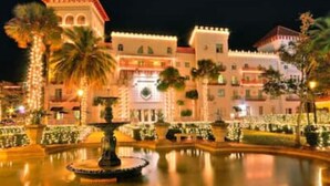 Un grand hôtel illuminé, doté d’une fontaine centrale, entouré de jardins bien entretenus et de palmiers.