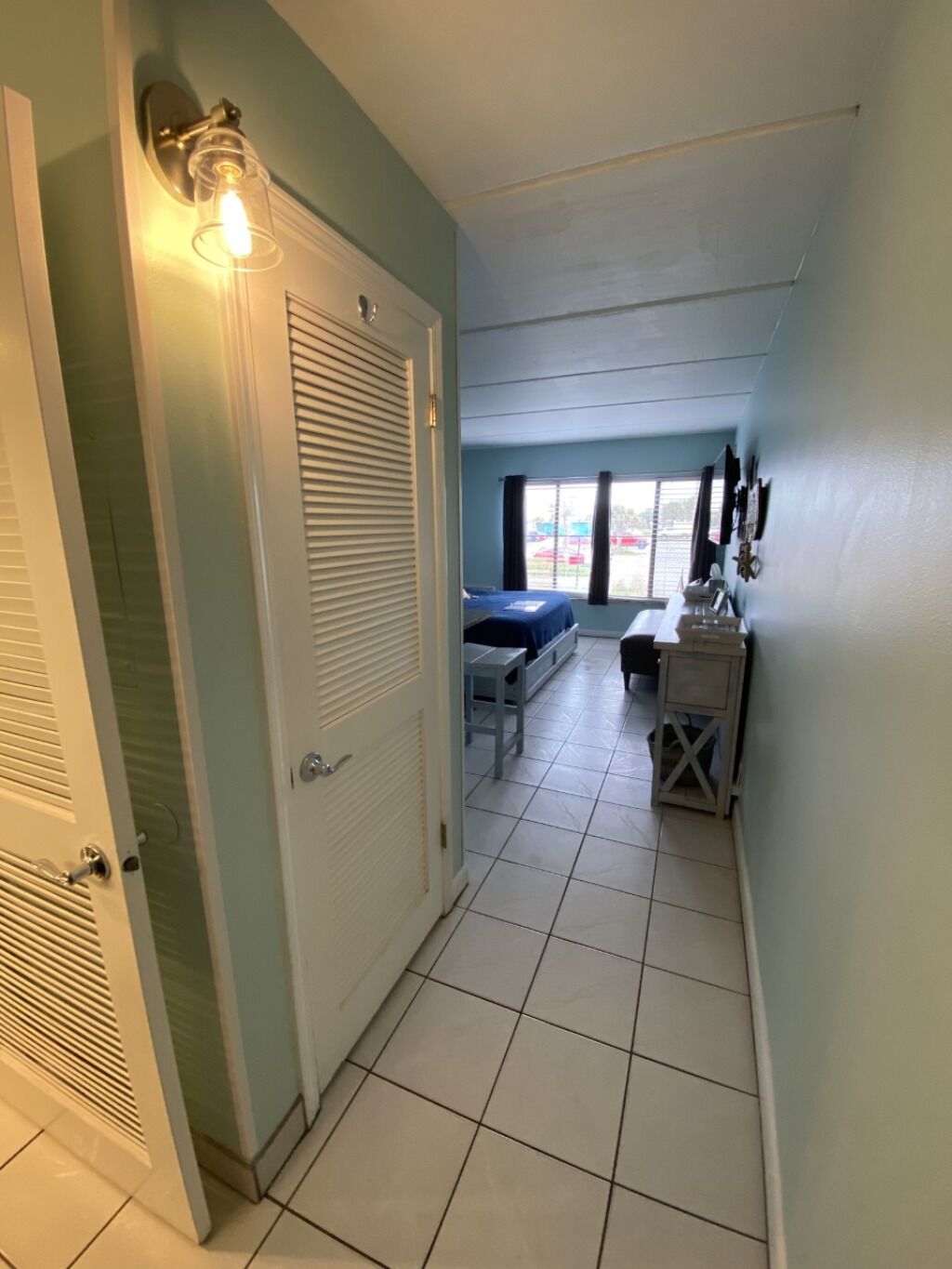 A hallway leading to a bedroom with a bed, a desk, and a window.