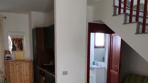 Ampla sala de estar com piso em azulejos, um sofá verde, um armário de madeira e uma escada com corrimão vermelho.