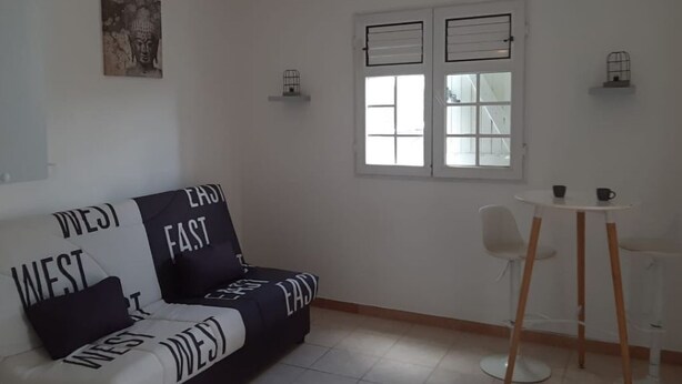 A modern living room with a sofa labeled "WEST" and "FAST," a dining area with a small table and chairs, and a window with blinds. - Charming, fully-equipped studio with mountain and sea views, 5 minutes from the beach (Schoelcher)