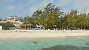 Ein Strand mit weißen Liegestühlen und Sonnenschirmen, ein Haus im Hintergrund und klares türkisblaues Wasser.