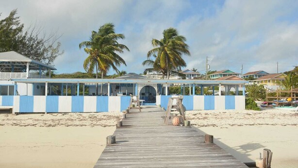Ein Steg am Strand, der zu einem Gebäude mit blau-weißer Fassade führt, mit Palmen und Strandhäusern im Hintergrund.