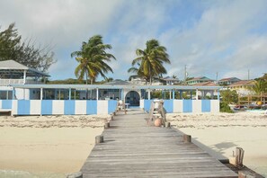 Een pier aan het strand die leidt naar een gebouw met een blauw-wit gevel, met palmbomen en strandhuisjes op de achtergrond.