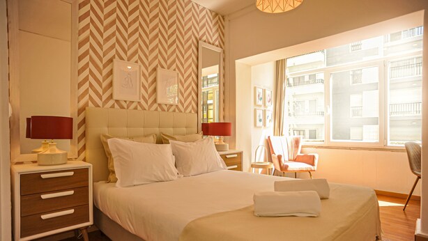 Een hotelkamer met een groot bed, twee nachtkastjes, een stoel en een raam met uitzicht op gebouwen.