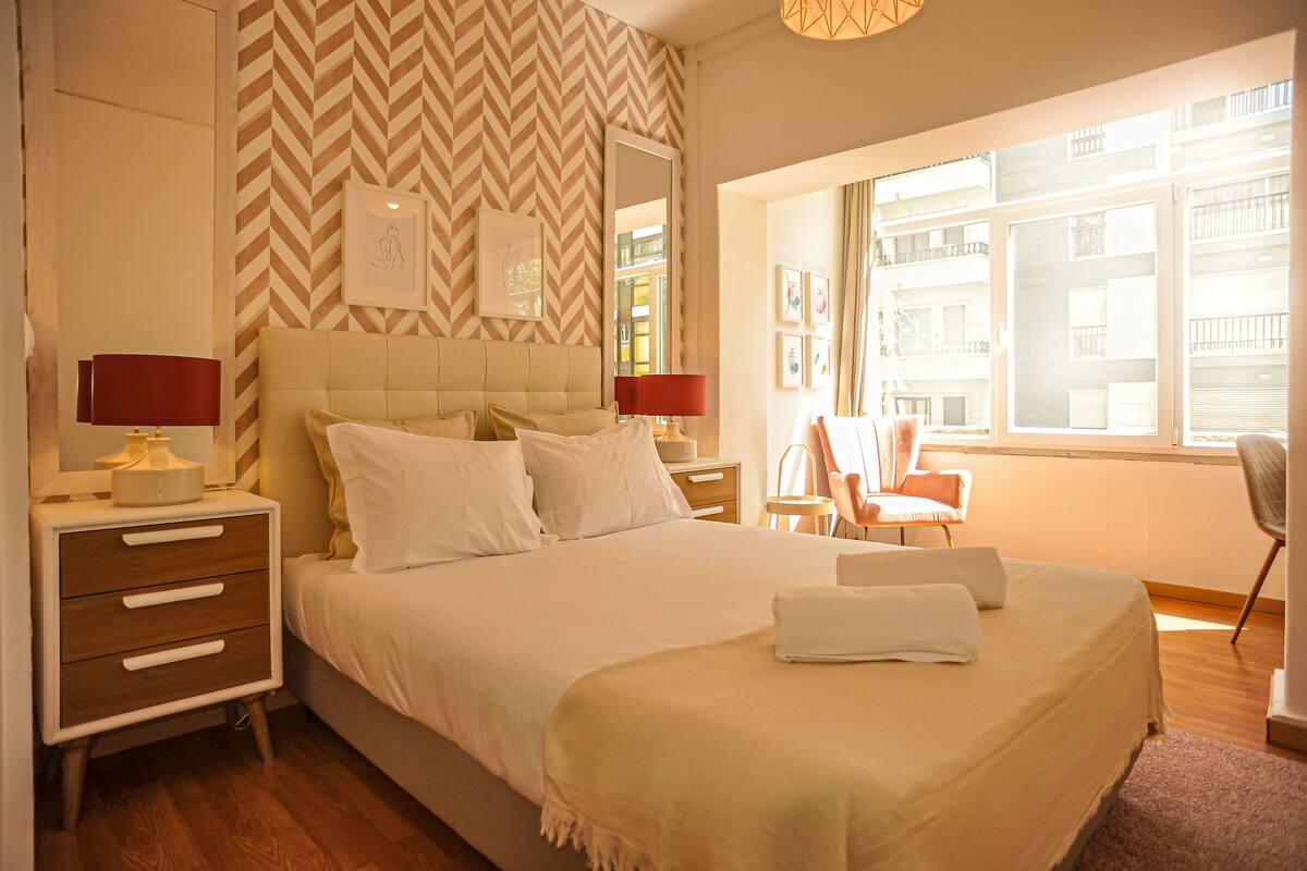 Une chambre d’hôtel avec un grand lit, deux tables de chevet, une chaise et une fenêtre donnant sur des immeubles.