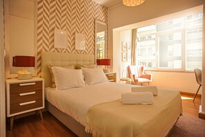Habitación de hotel con una cama grande, dos mesitas de noche, una silla y ventana con vistas a edificios.