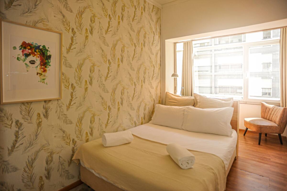 Une chambre d’hôtel avec un lit, une chaise, une fenêtre et un mur orné d’un motif floral.