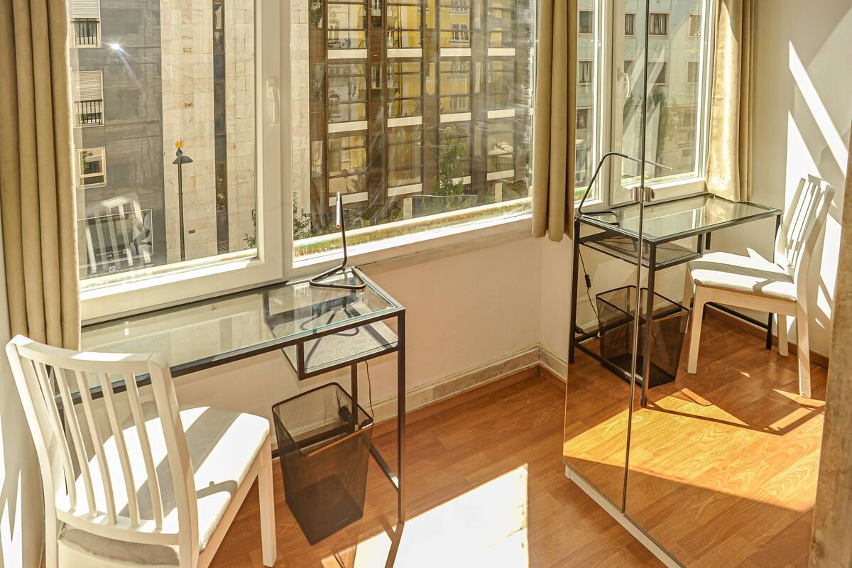 Une pièce avec un bureau en verre, deux chaises blanches et une vue sur le paysage urbain, visible à travers de grandes fenêtres.