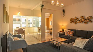 Een moderne woonkamer met een bank, fauteuil en salontafel. Er is een eethoek met een tafel en stoelen. De kamer heeft een groot raam en uitzicht op buiten.