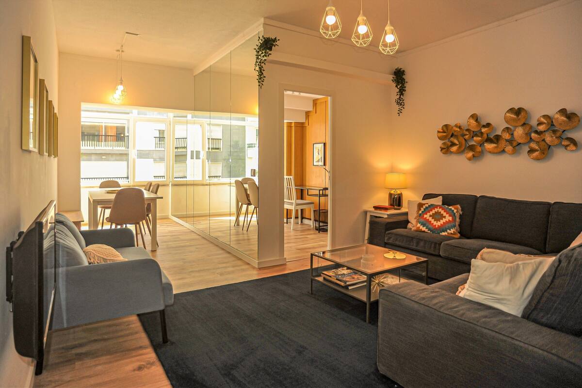 Un salon moderne avec un canapé, un fauteuil et une table basse. On y trouve également un coin repas avec une table et des chaises. La pièce est dotée d’une grande fenêtre qui offre une vue sur l’extérieur.