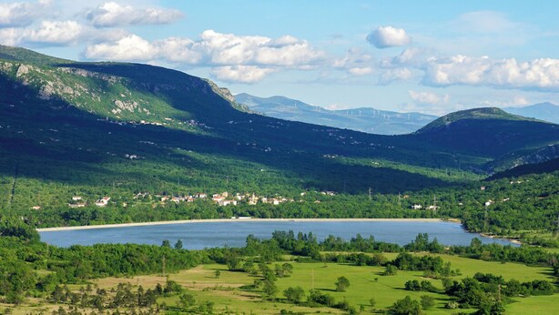 Une vue panoramique d’un lac entouré de collines verdoyantes et d’une petite ville dont on aperçoit les bâtiments.
