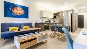 A modern living room with a blue sofa, wooden coffee table, and a painting of a turtle. Adjacent is a kitchen with wooden cabinets, a refrigerator, and a dining area with chairs. - Veranto (Playa del Carmen)