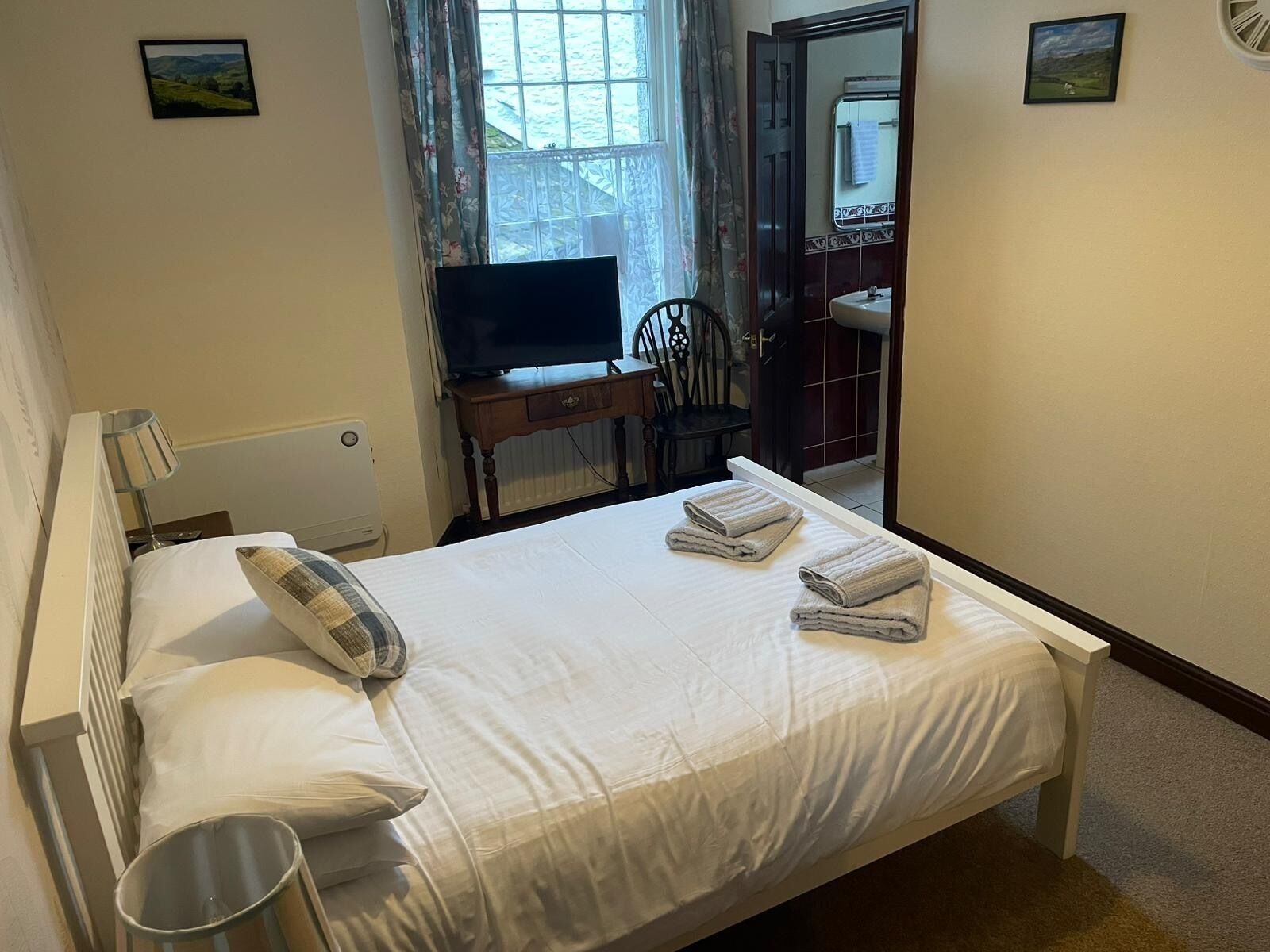 A neatly made bed with pillows, a bedside lamp, a TV on a wooden stand, and a bathroom visible through an open door.