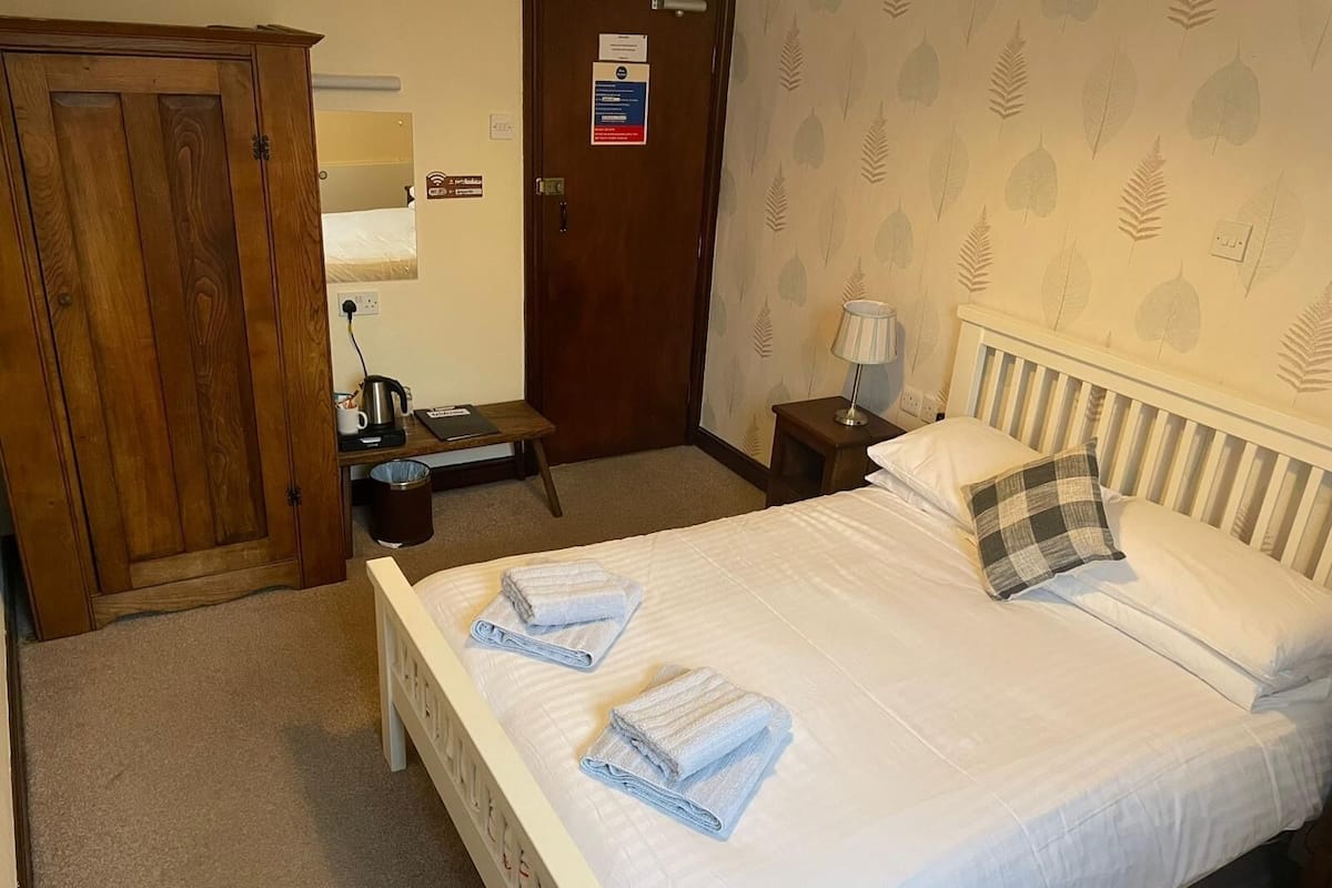 A hotel room with a bed, a wooden wardrobe, a small table with a kettle, a lamp, and a wall with leaf-patterned wallpaper.