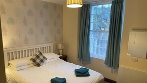 A neatly made bed with pillows, a bedside lamp, a window with curtains, and a radiator. - George and Dragon Dent (Sedbergh)