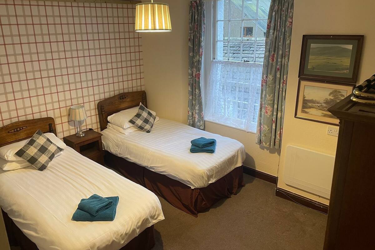 A hotel room with two beds, a window with curtains, a lamp, and a desk with a telephone.